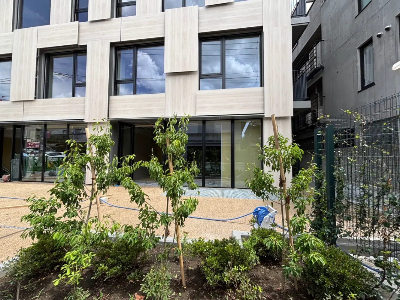 - 近代的な建物の外観、ガラス窓と木製パネルのファサード、周囲に植えられた若木と庭のデザイン