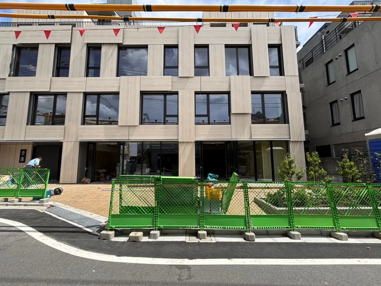 - 現代的なデザインの建物、ガラス張りのファサードと植栽、工事用の緑のフェンスが設置された外観