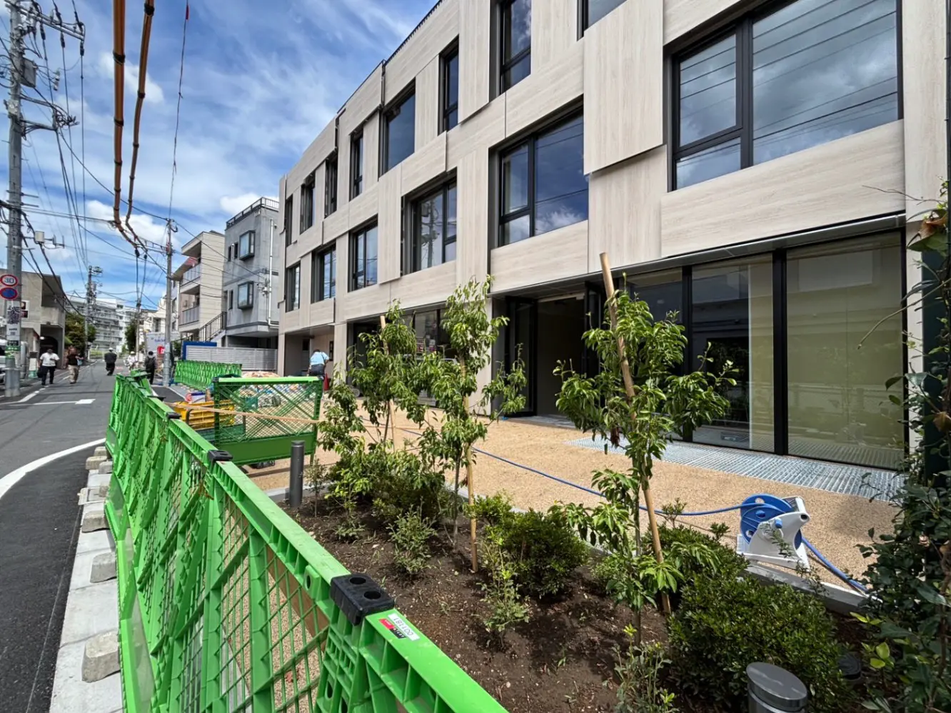 - 現代的な外観の建物、緑のフェンスに囲まれた庭と舗装された通路、周囲を歩く人々が写った都市風景