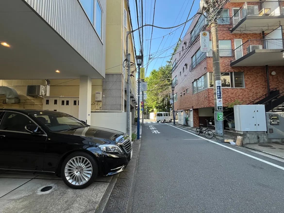 - 黒い高級車が停車している都市の街並み、周囲にはアパートや電柱、青空が広がる風景