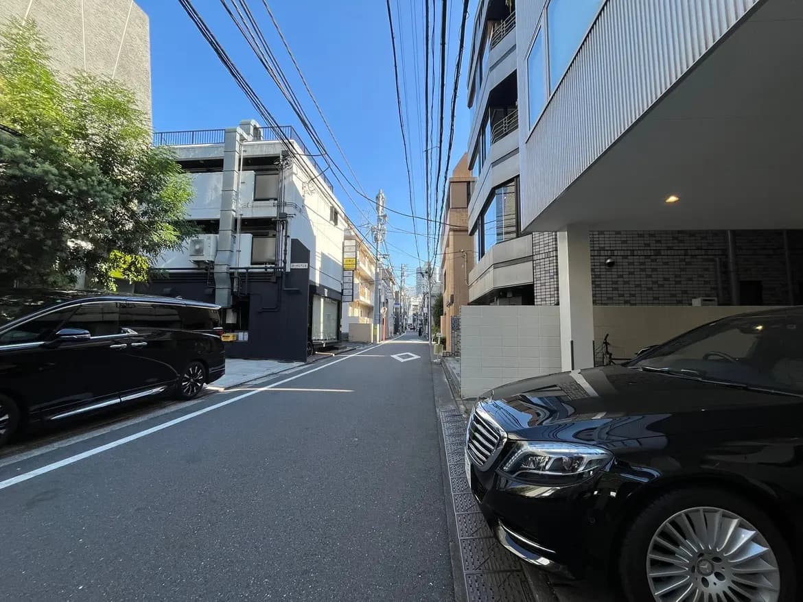 - 青空の下、道路と並ぶマンション、停車中の黒い車と電線が見える都市の風景