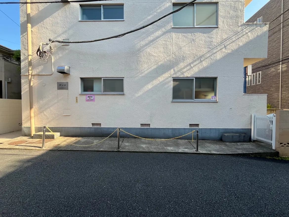 - 白い外壁の建物、窓が二つあり、正面に「クンシ」の名前が掲示されている、周囲には舗装された道路が広がる景観