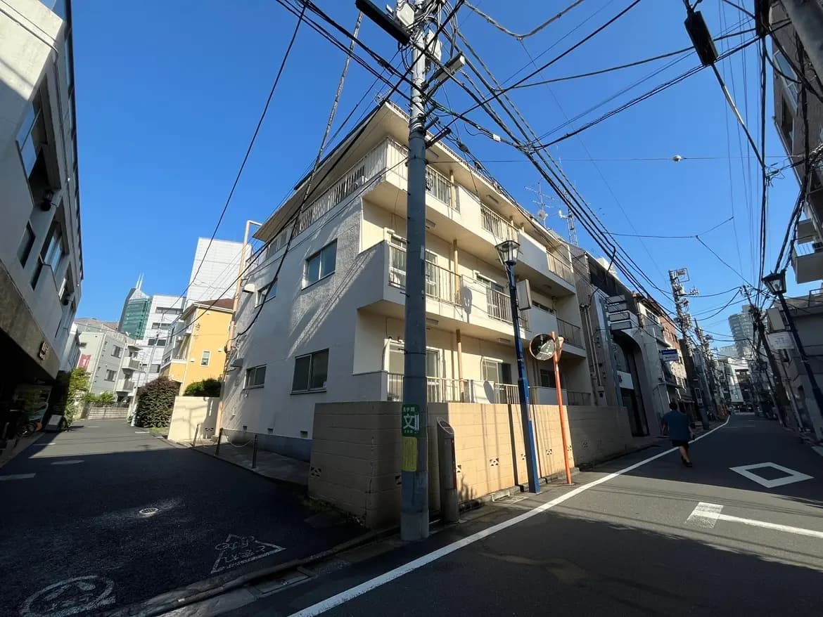 - 青空の下に広がる街角、マンションと電柱、多様な建物が立ち並ぶ風景、右手には高層ビルが見える通りの様子