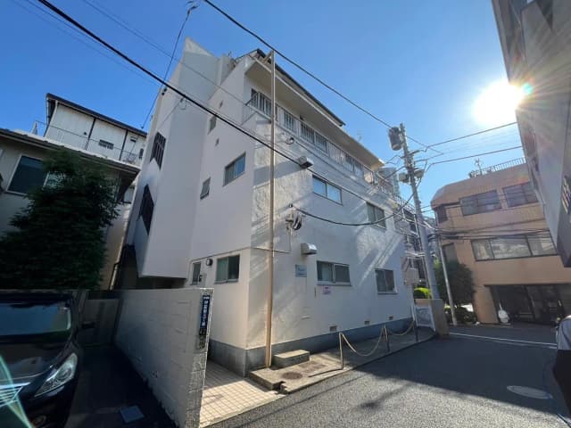 - 青空の下、白い外壁の集合住宅と隣接する建物、道路の電線と停車中の車が見える都市の風景