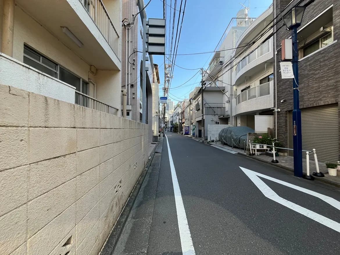 - 住宅街の細い道路、左右に並ぶ建物と青空が広がる風景、電柱と配線が見える通りの様子