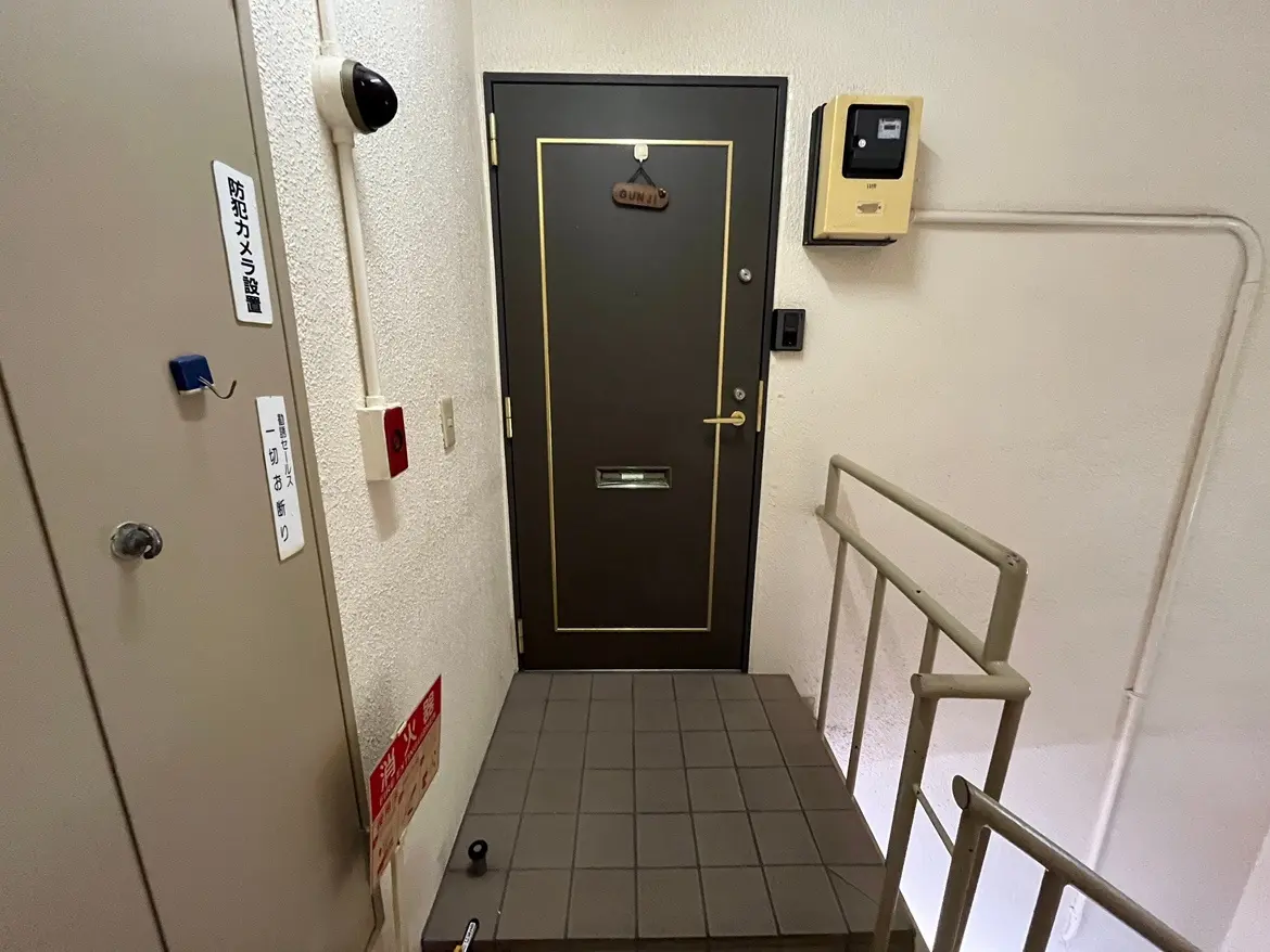 - 防犯カメラが設置されたエントランスのドア、黒いドアに金のライン、近くに郵便受けと案内表示がある玄関ホール