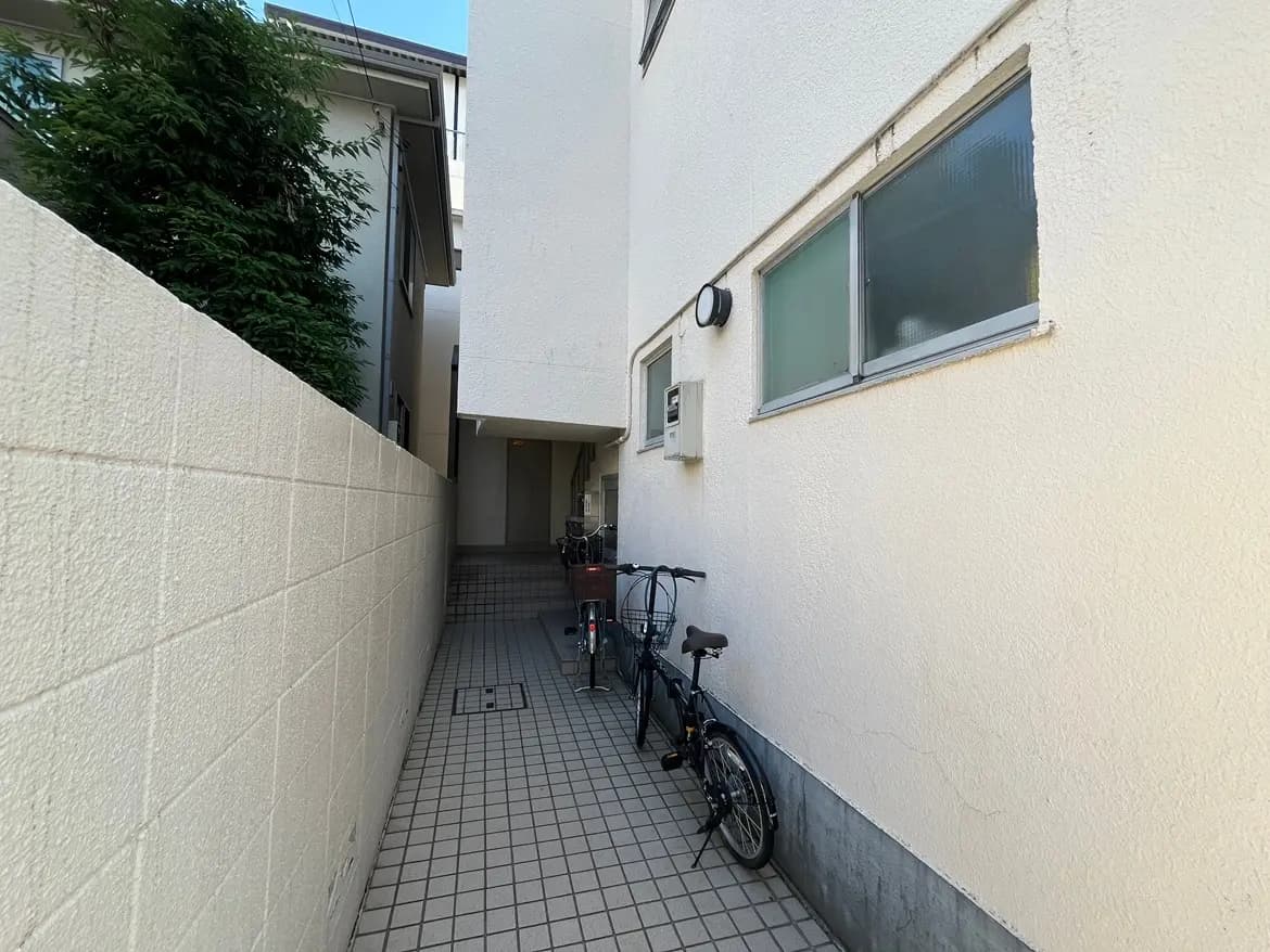 - 住宅の横にある狭い通路、白い壁とタイルの床、停められた自転車と緑の植物がある風景