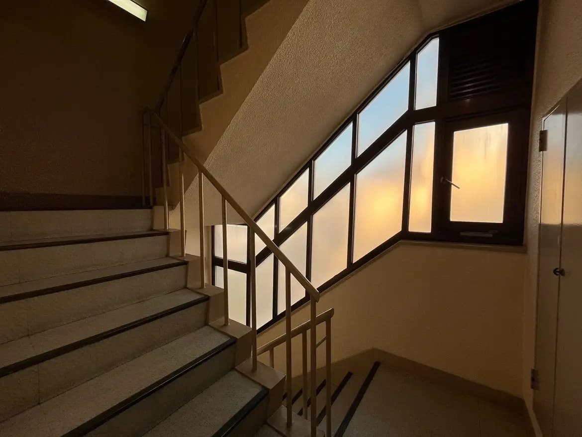- 階段の手すりと明るい窓がある暗い色調の階段室、窓からは夕焼けの光が透けて見える