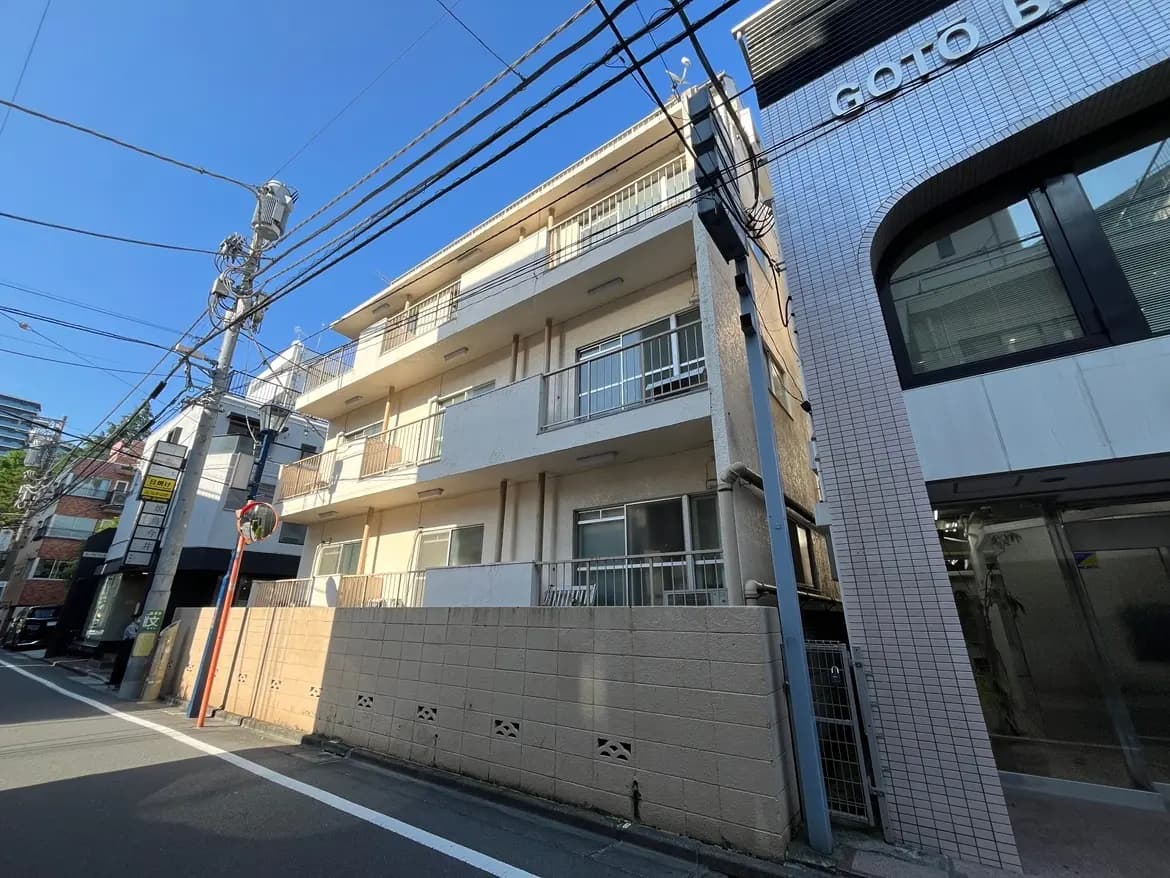 - 道路に面した古いアパートの外観、隣にモダンなビルが立ち並ぶ、青空と電線が見える景観