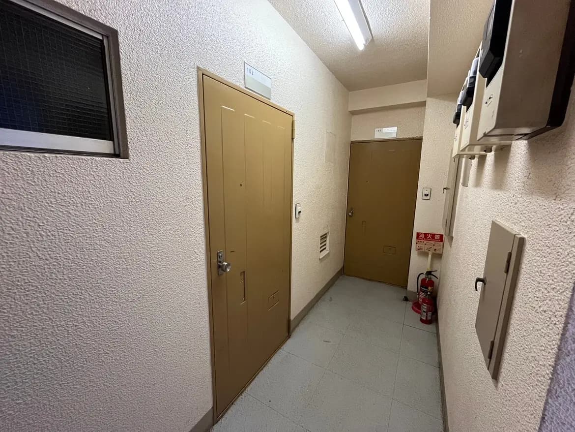 - シンプルなデザインのアパートの廊下、薄い壁と黄褐色のドアが並ぶ静かな空間、壁際に設置された火災警報器と消火器を含む安全設備