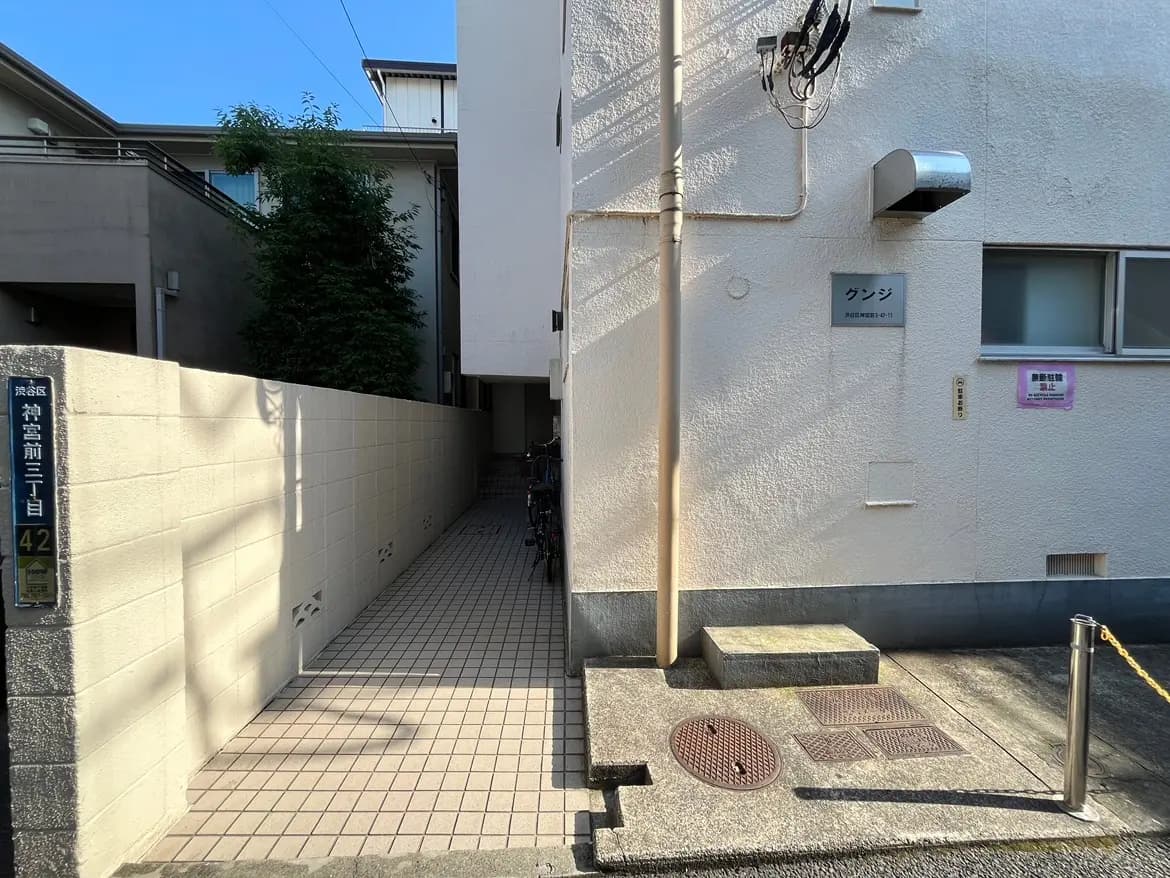 - 立て看板のある白い壁に囲まれた住宅の通路、右側に自転車が並ぶ風景と青空の背景