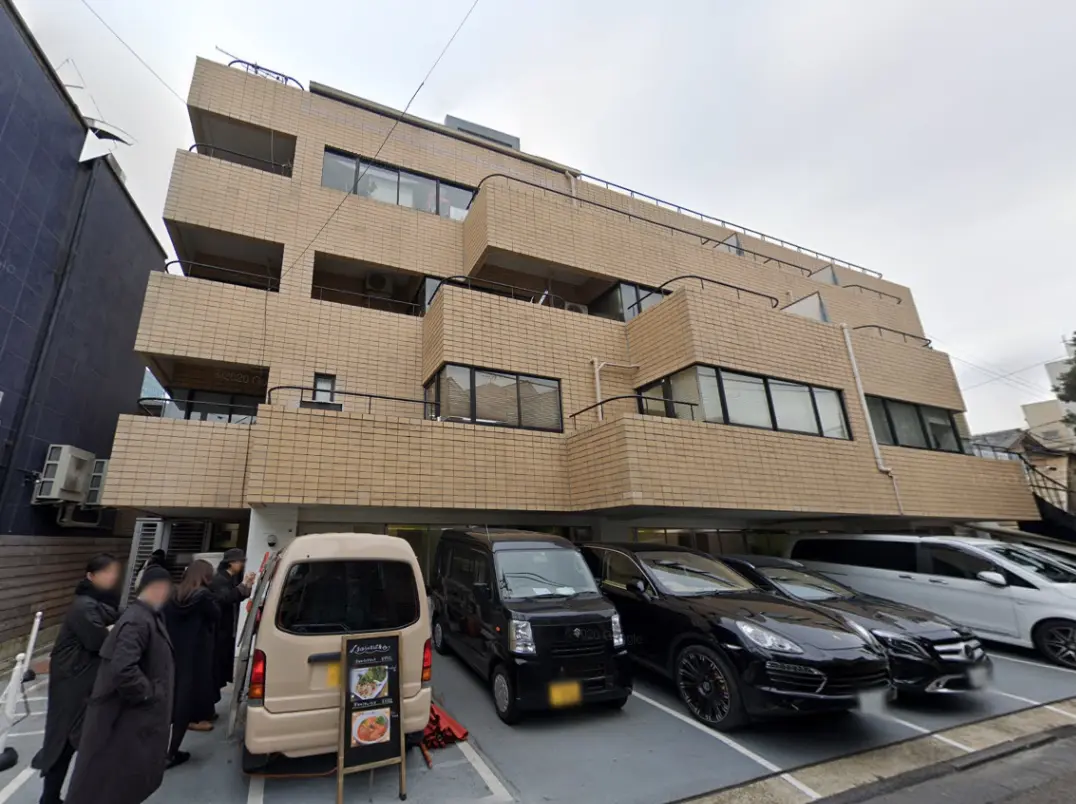 - 若干モダンな外観のアパートメントビル、駐車場には複数の車があり、食事メニューの看板が目立つシーン