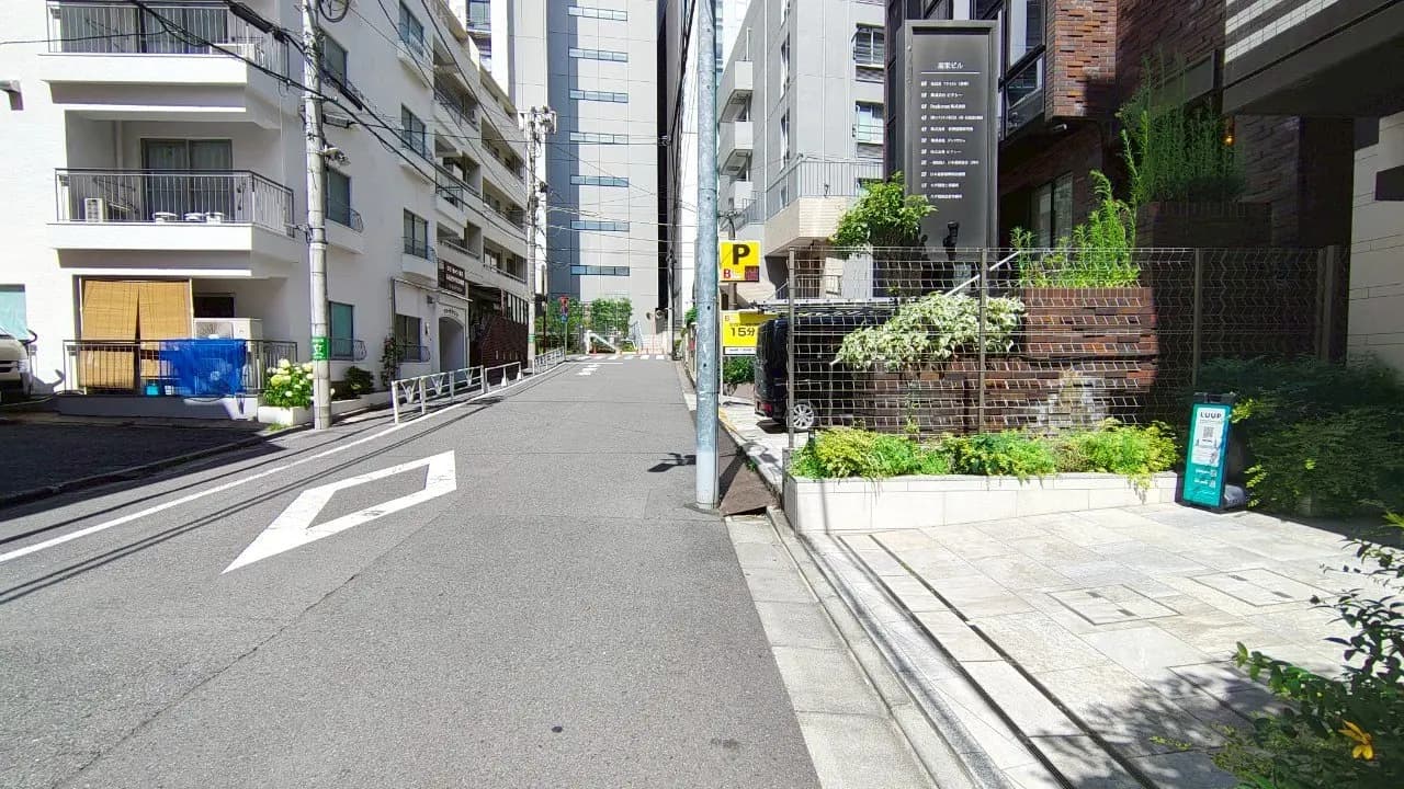 - 都会的な住宅街の風景、左側にバルコニーのある建物、右側には緑豊かな植栽と看板が見える通りの様子
