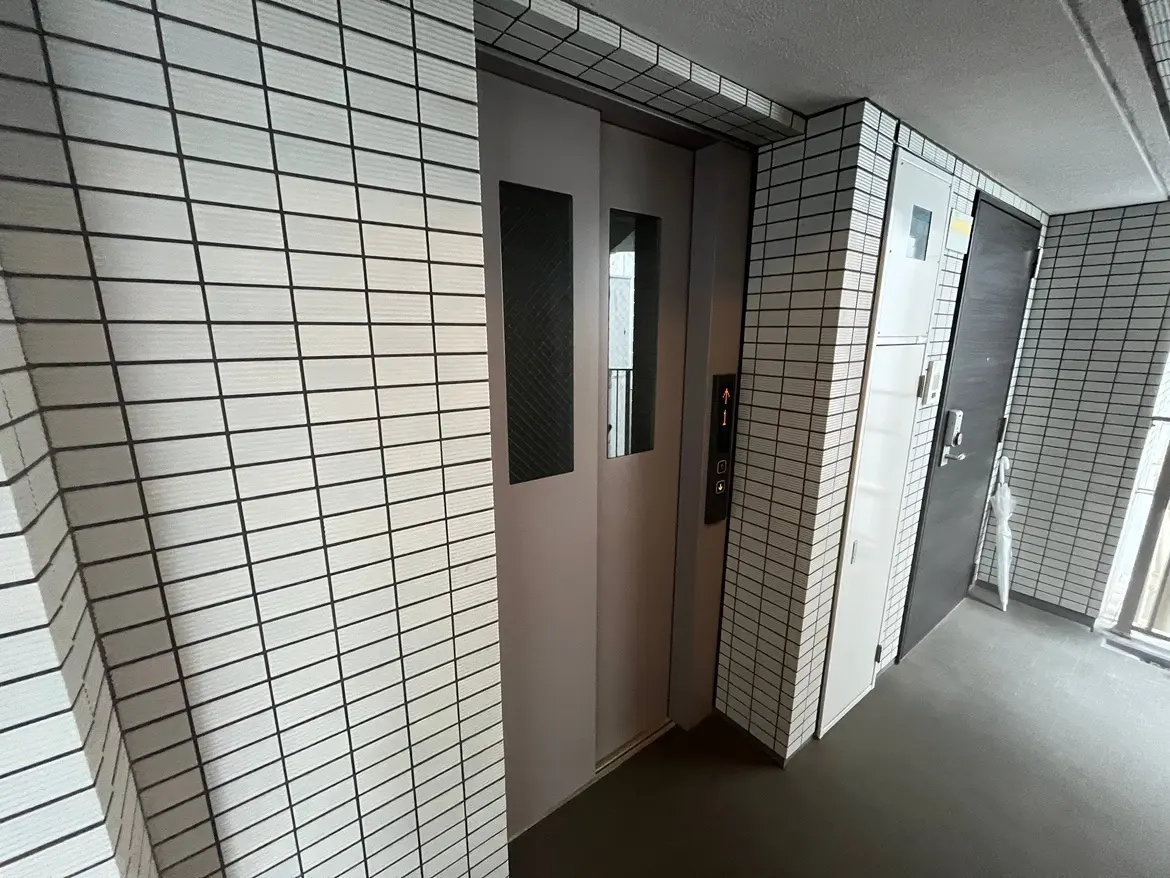 - シンプルでモダンなエレベーターの入り口、タイル張りの壁と隣接するダークカラーのドアが特徴の廊下の風景