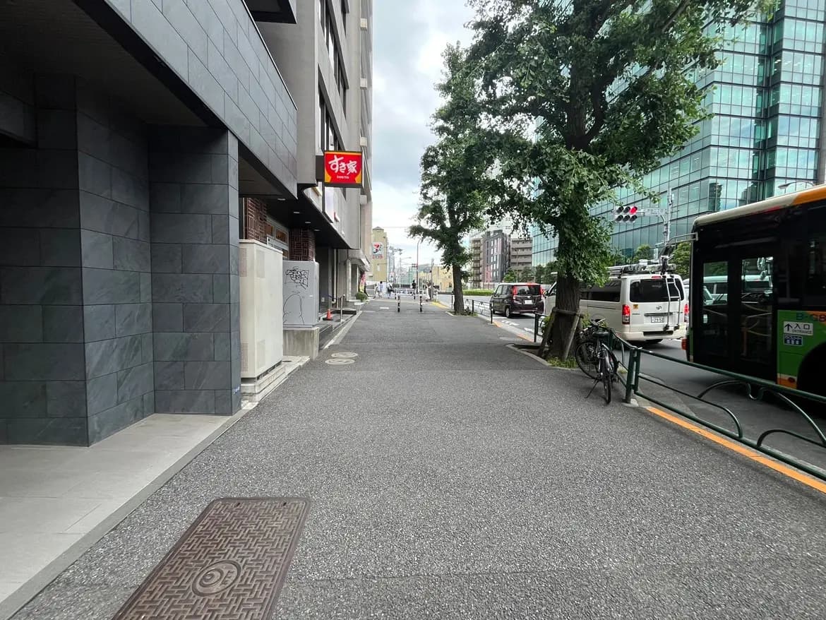 - 商業ビルの隣にある歩道、黒いタイル張りの壁、背景には緑の木々とビル群が見える街の風景