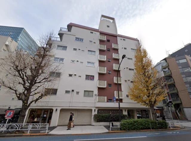 - 都市型のアパートメントビル、クリーム色の外壁に赤褐色の部分、周囲に並ぶ樹木と人々が通行する様子
