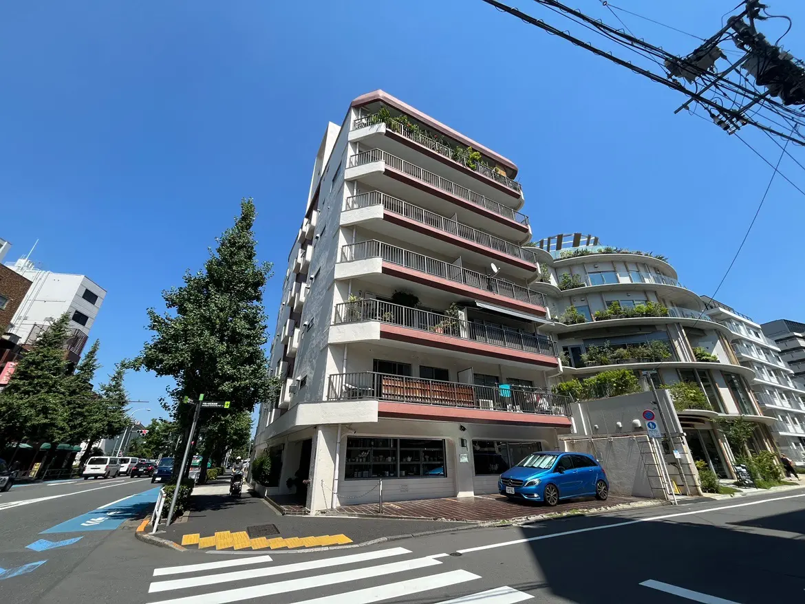 - 青空の下にある現代的なマンションの外観、周囲の緑と交通量の多い街角が特徴、道路には駐車中の車も見える