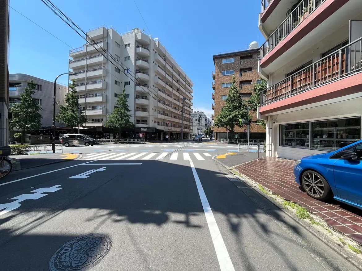 - 明るい青空の下、交差点と周囲のビル、道路に描かれた白い横断歩道と駐車中の青い車が見えるシーン