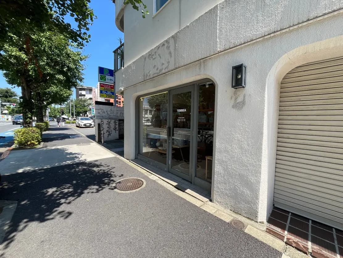 - 明るい日差しの下、白い壁の店舗入口と近くの街路樹、周辺には車と歩行者の様子が見える風景