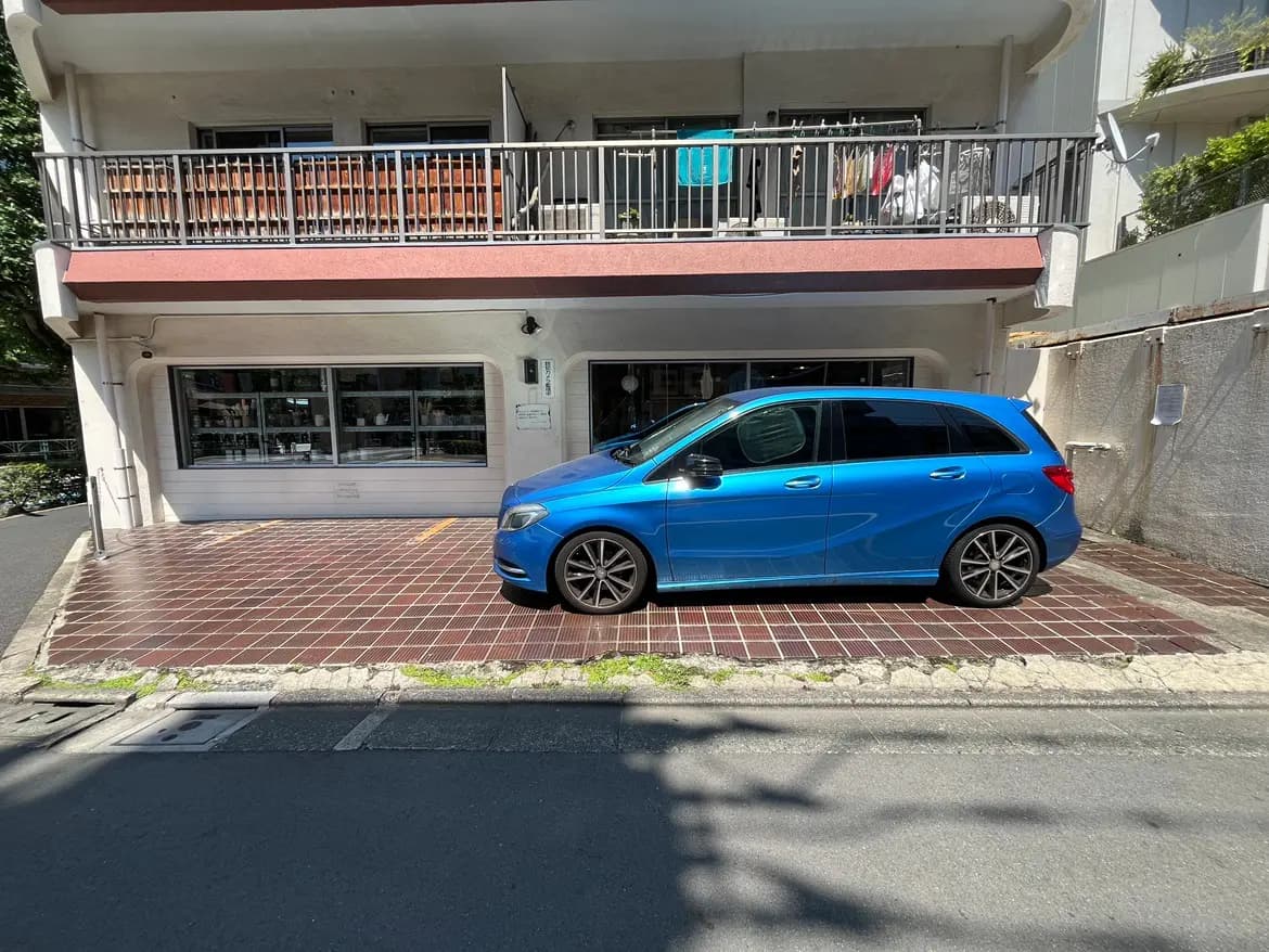青いコンパクトカーが停まっているマンションの前景、晴れた日の明るい室外、壁に洗濯物が干されている様子