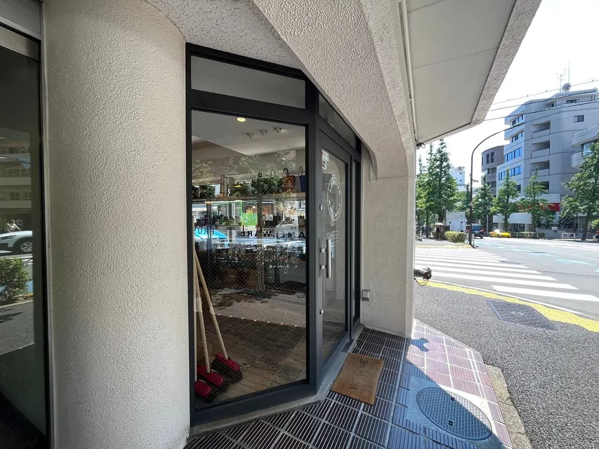- 店舗の入り口、ガラスのショーウィンドウと白い壁、周囲に歩道と緑の木々がある街並み