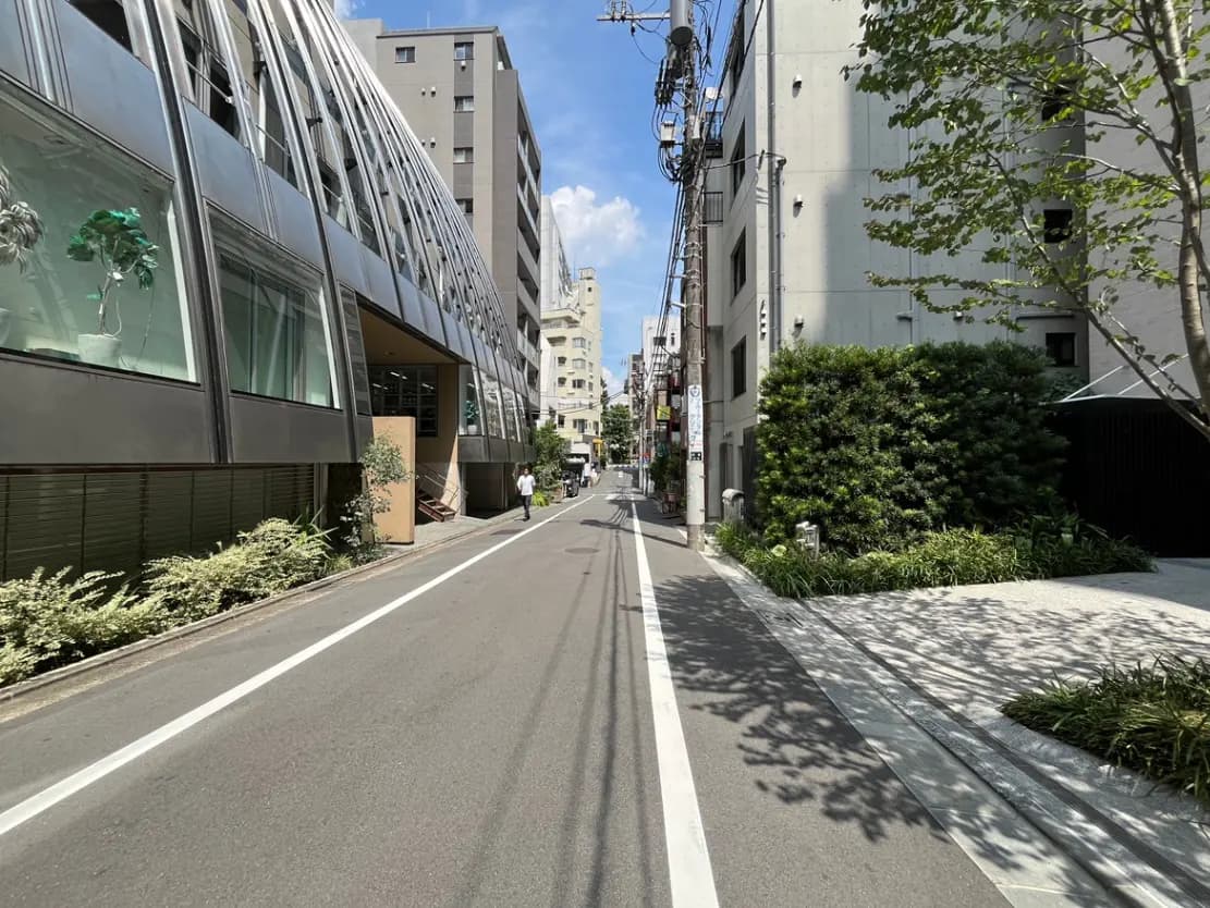 - 都会的な雰囲気のある通り沿いの風景、左側に近代的な建物、道を歩く人と緑の植栽が特徴的なシーン