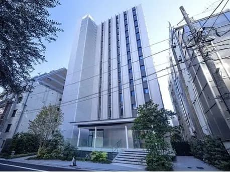 - モダンなデザインの高層ビル、透明なエントランスと周囲の緑豊かな植栽、都会的な景観の中に立つ建物