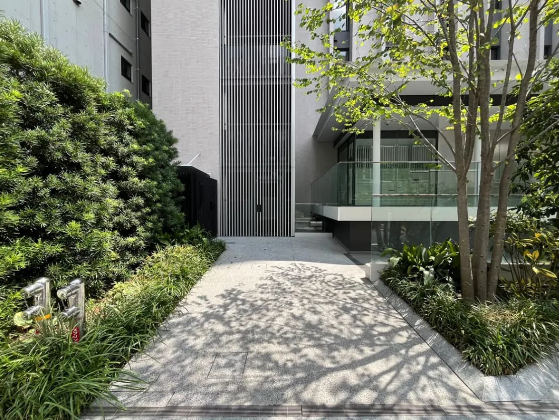 - モダンな建物のエントランス、緑豊かな植栽とシャープなデザインの外観、光が差し込む歩道が広がるエリア