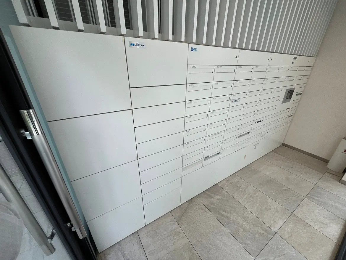 - 白を基調とした郵便受けボックス、各部屋番号が記された配達用ロッカーが整然と配置されたエントランスの内部景観