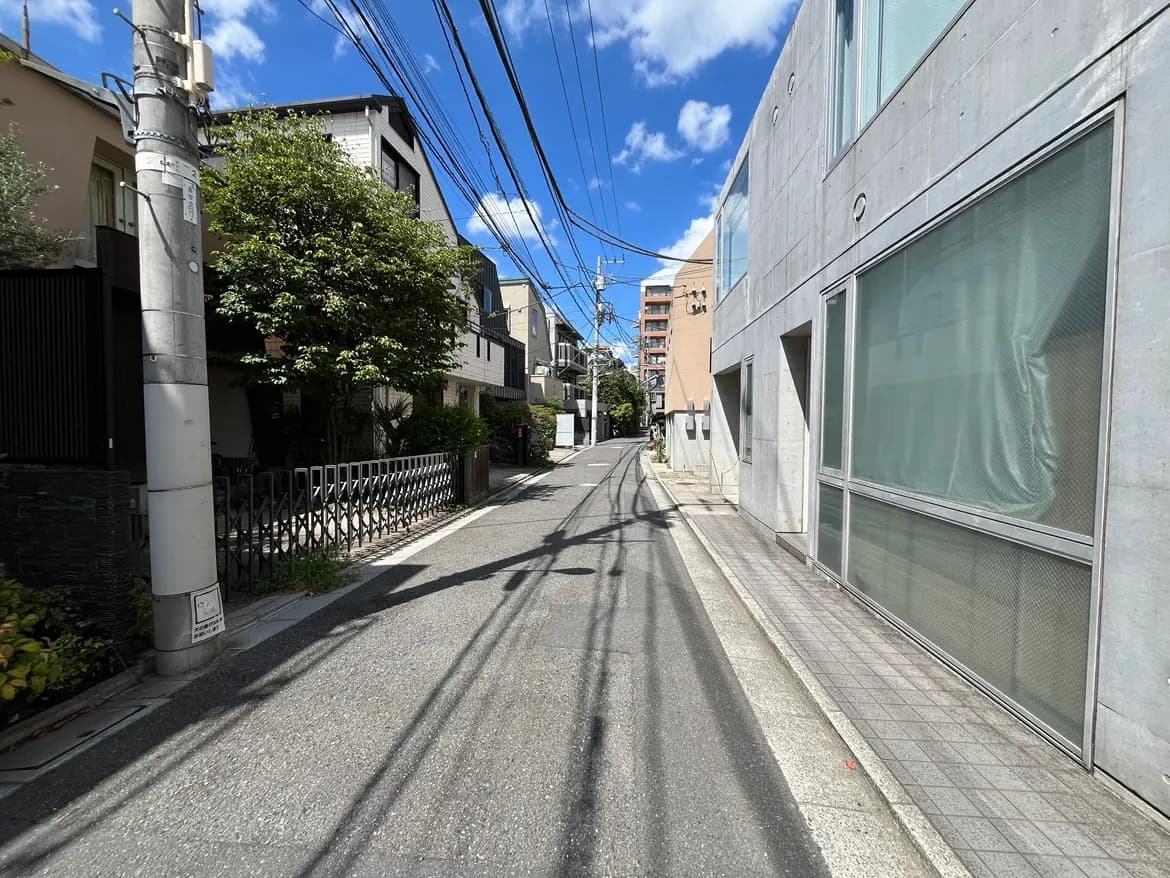 - 青空とおしゃれな建物が並ぶ街並み、左側に植栽された街路樹と電線が見える静かな通りの風景