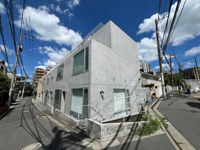 - コンクリート外壁のモダンな建物、青空に浮かぶ白い雲、周囲の電線と狭い道路が見える都市景観