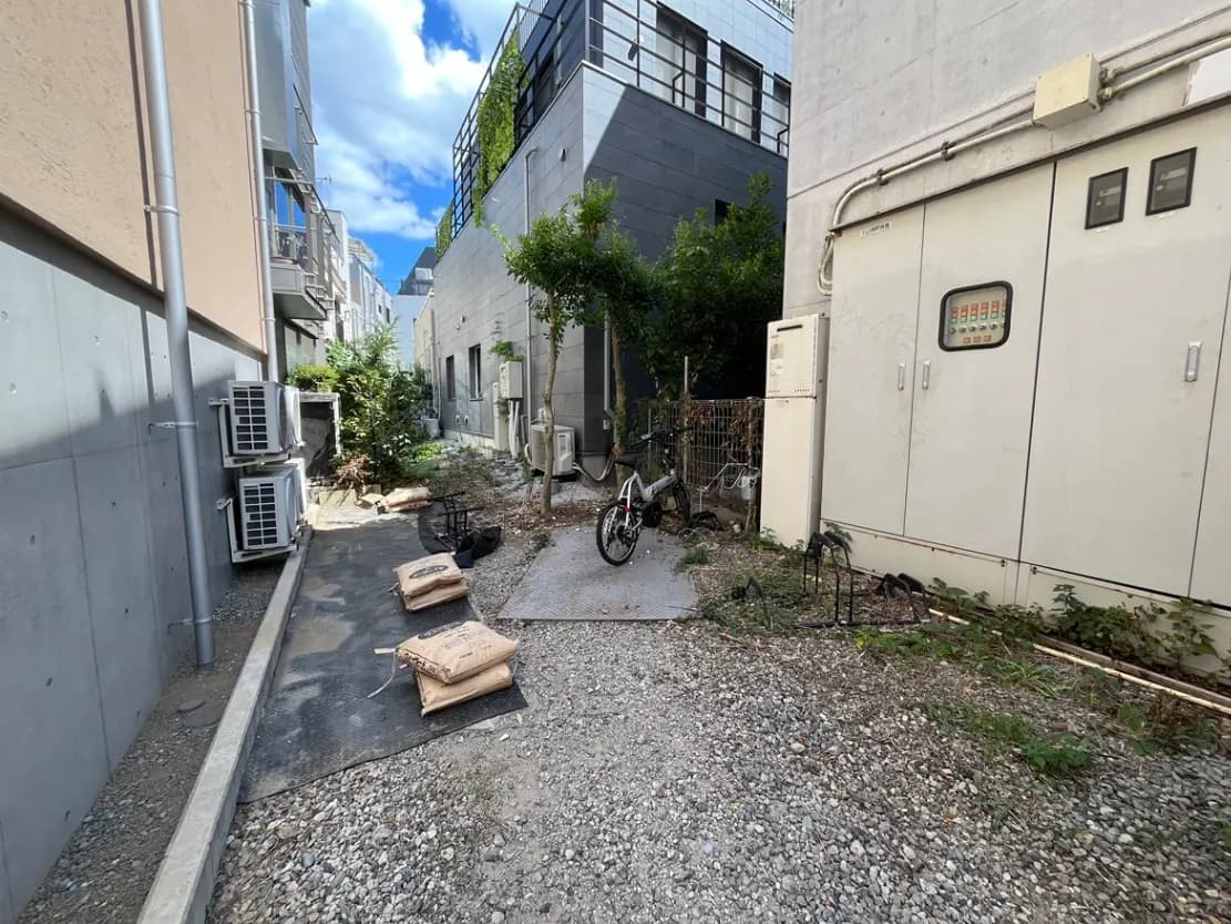 - 狭い裏通りの風景、隣接した建物、エアコンの室外機、雑草が生えた地面と自転車がある場所