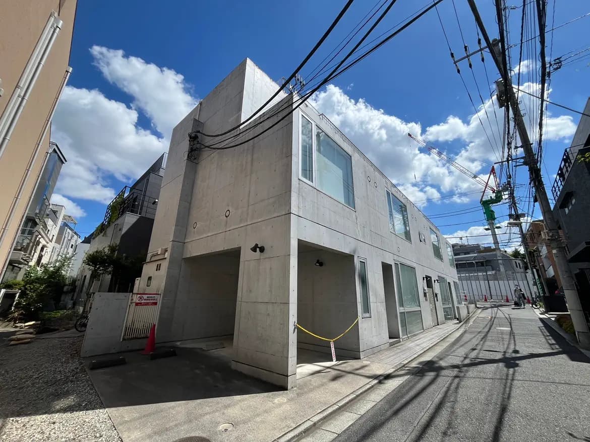 - コンクリート外壁のモダンな建物、青空と白い雲が広がる日中の風景、周囲の建物や電線が視界に入る街並み