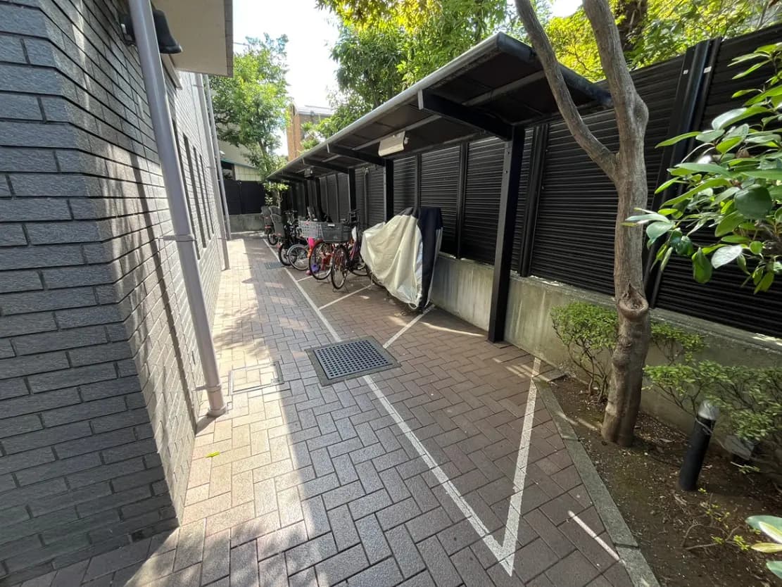 - 自転車置き場の通路、物置きと自転車が並んでいる、周囲は緑の木々に囲まれたエリア