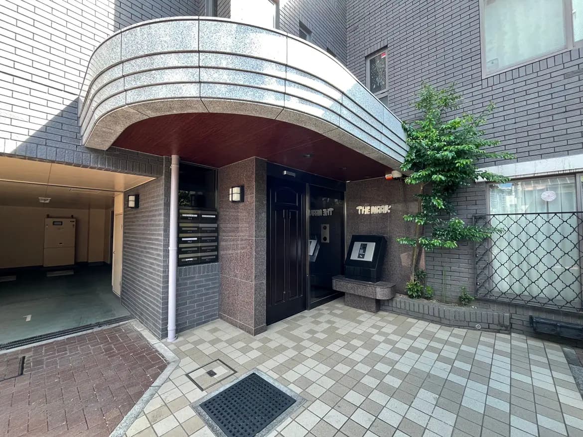 - モダンなアパートのエントランス、グレーのタイル張りの壁、木製のバルコニー、郵便受け、周囲に緑の植物がある外観