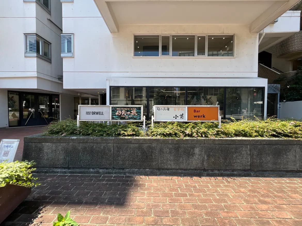 - 店舗の外観、様々な看板が並び、周囲には緑の植栽とレンガ敷きの道がある光景
