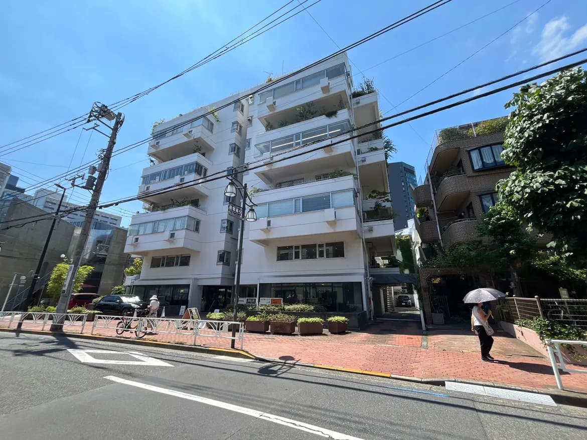 - 現代的なデザインのマンション、青空の下で白い外壁とバルコニーの緑が映える街並み、近くに自転車を押しながら歩く人と植栽された歩道がある景観