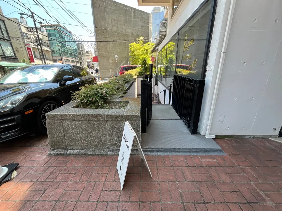- 店舗の入口前に置かれた案内看板と、植栽が施された石造りの花壇が見える街並みの風景。