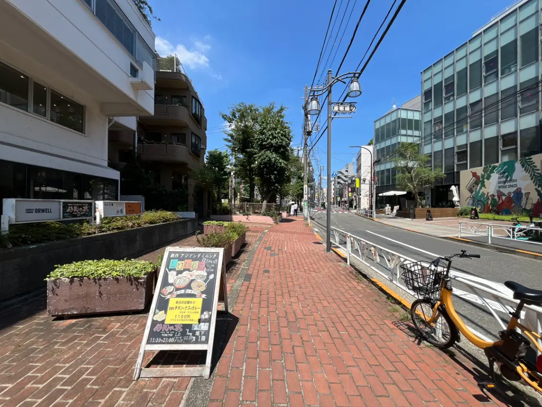 - 明るい青空の下、レンガ道沿いに並ぶビルや草花、看板が置かれた歩道の風景、黄色い自転車が停まっている。