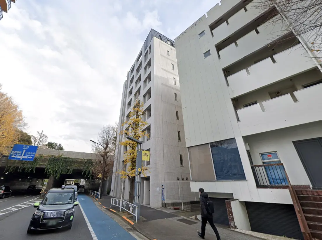 - 都市部に立地するモダンな建物の外観、青空と雲が広がる背景、歩道を歩く人と駐車中の車が描かれた風景