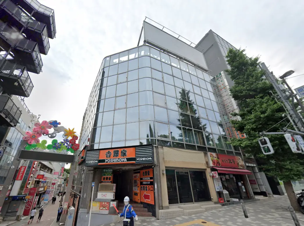 - ガラス張りのモダンなビルの外観、吉野家の看板と周囲の店舗、花のアーチが印象的な街並み