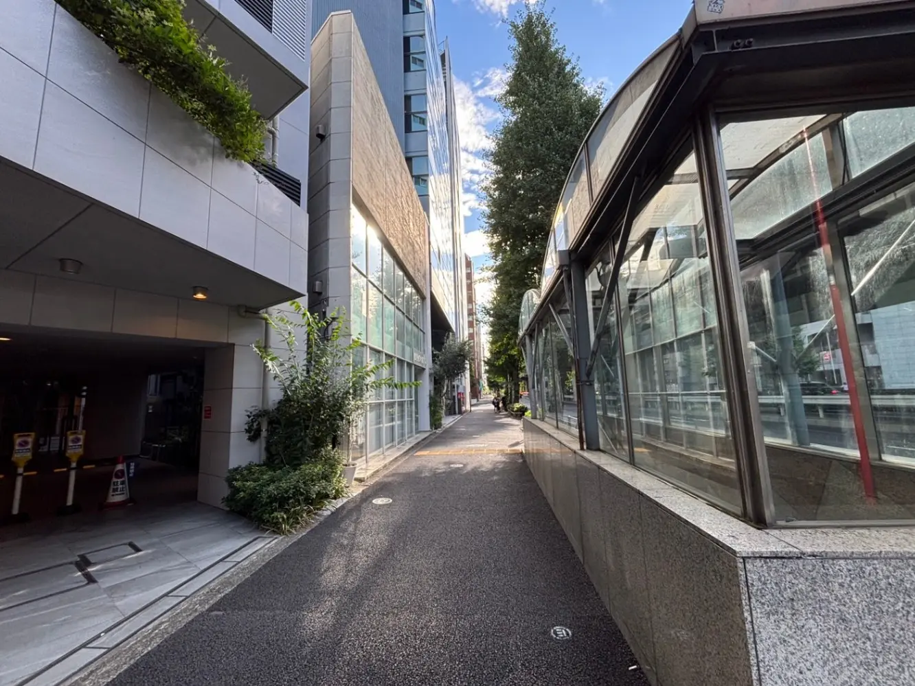 - 都会の歩道沿いの風景、近代的なビルとガラス張りのバス停、緑の植栽が点在する街並み