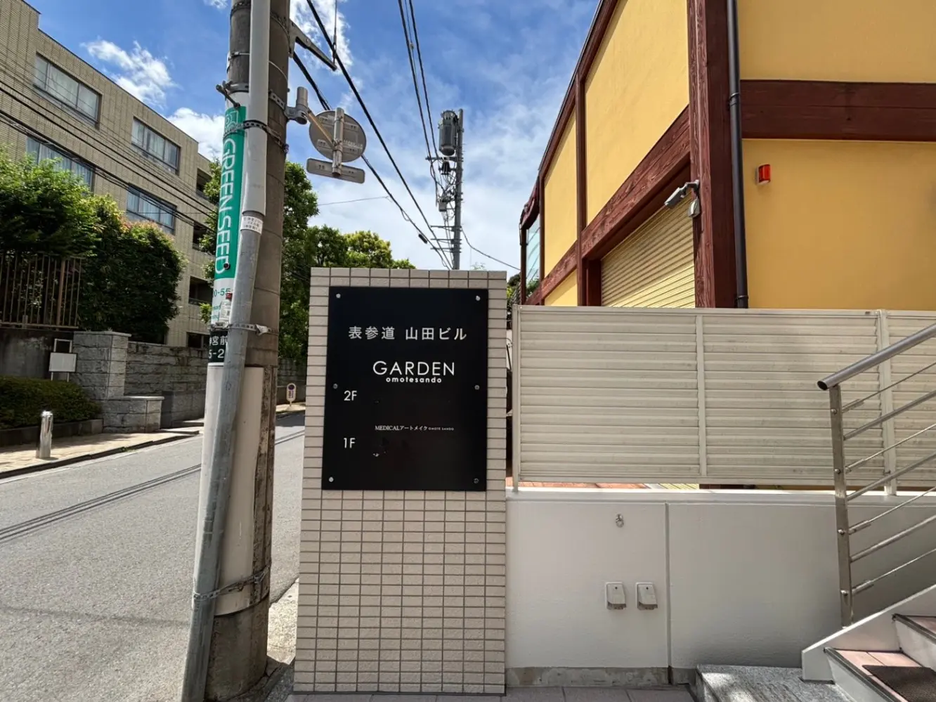 - 表参道の山田ビルの外観、階数案内と「GARDEN omotesando」の看板、周囲に緑と青空が広がる風景
