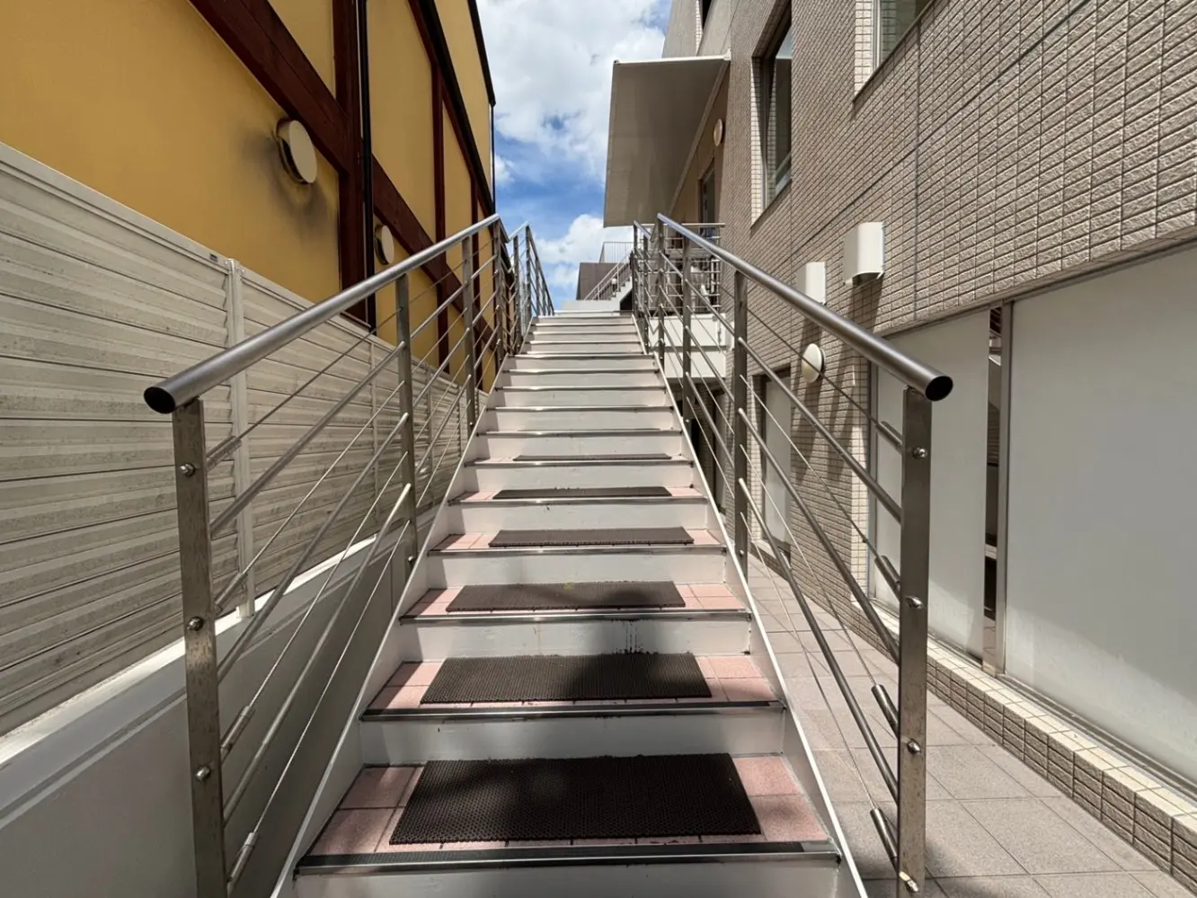 - 日差しが差し込む、金属製の手すり付きの階段、周囲にモダンな建物が立ち並ぶ風景