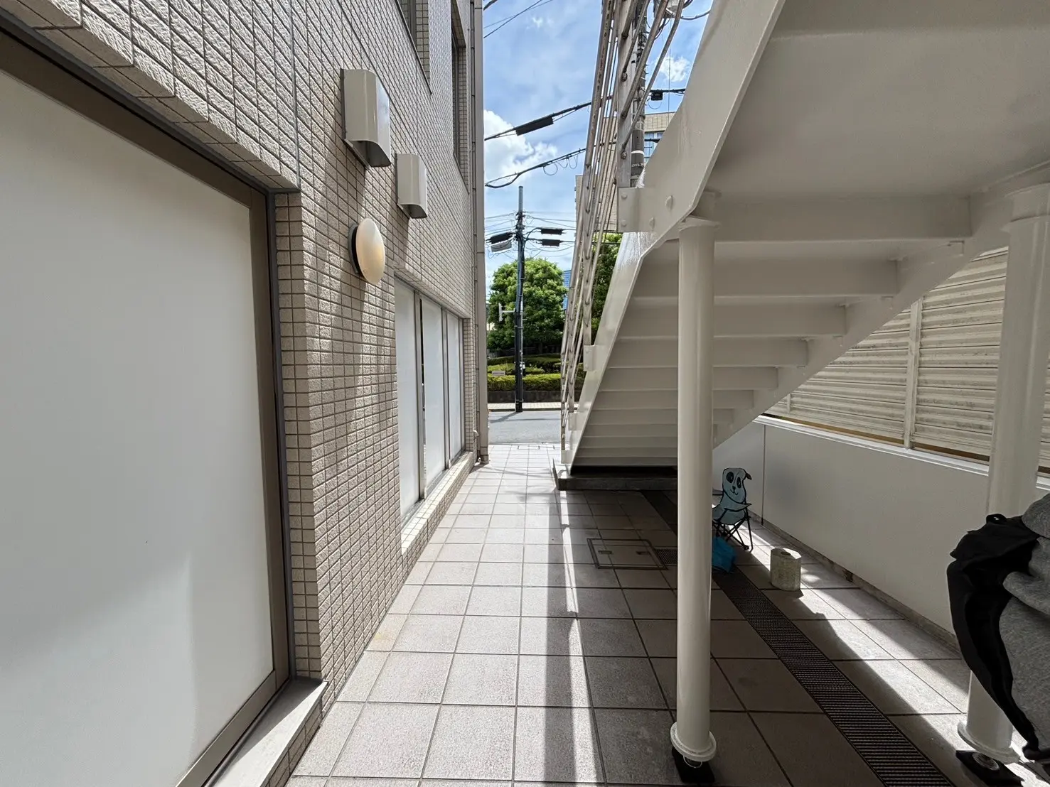 - 日差しが差し込む外階段とタイル張りの通路、隣接する建物の壁面と街路が見える風景