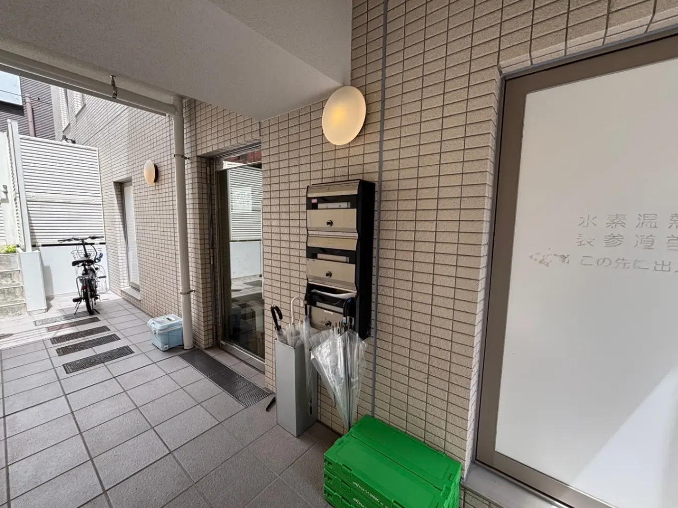 - グレーのタイル床が特徴のエントランスホール、郵便受けや傘立て、自転車が置かれたシンプルな空間