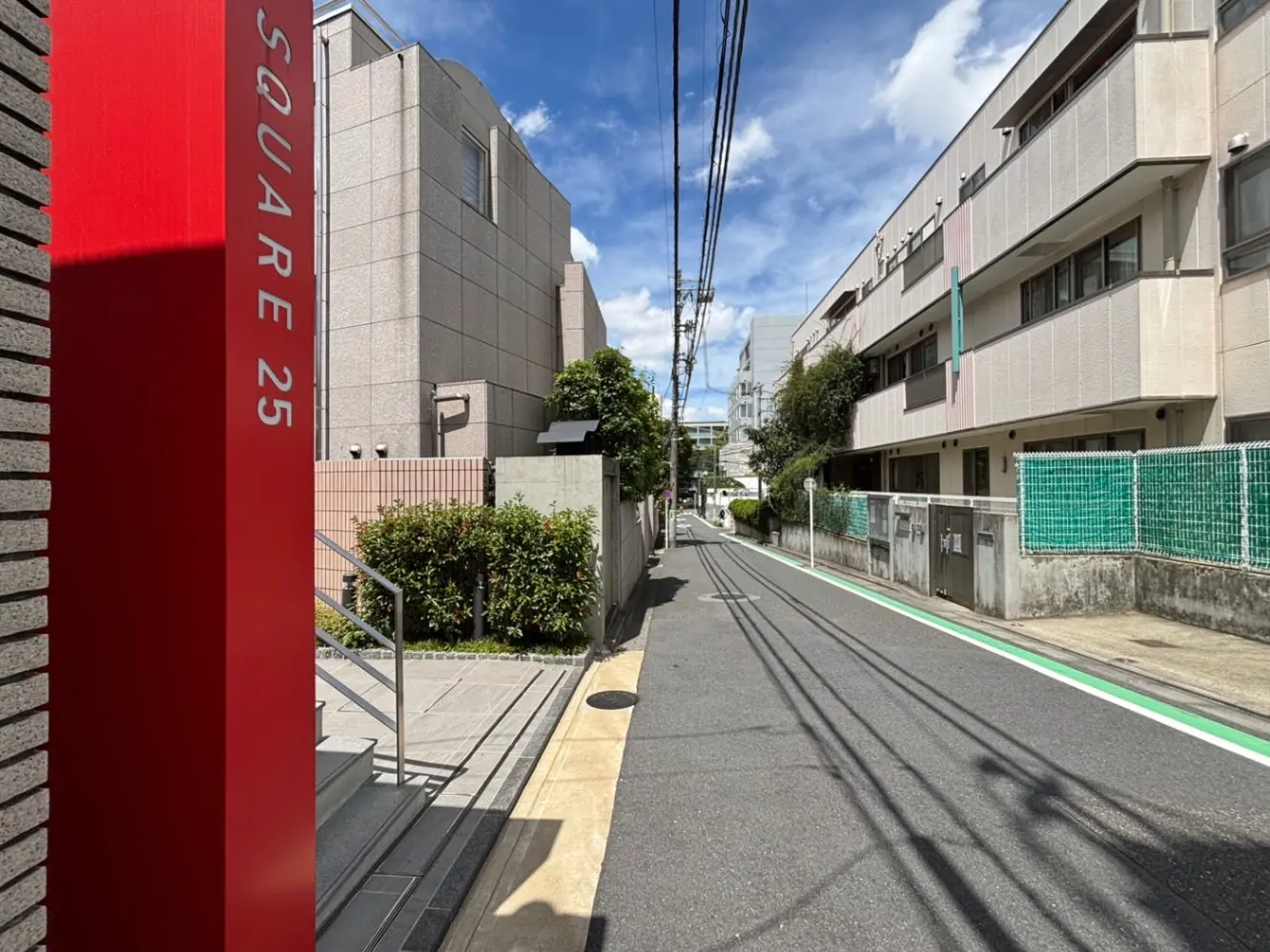 - 明るい青空の下、赤い看板「SQUARE 25」が目立つ住宅街の風景、周囲には緑の植物と近隣の建物が並ぶ通りの様子