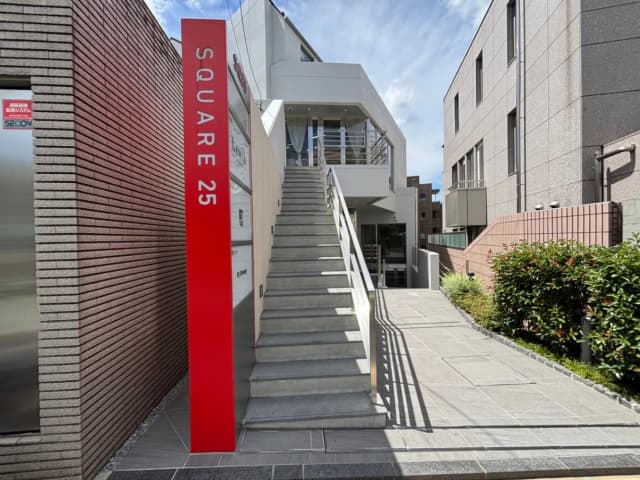 - 白い外観のビルのエントランス、階段と赤い看板「SQUARE 25」が目立つ現代的な建物の入り口の風景