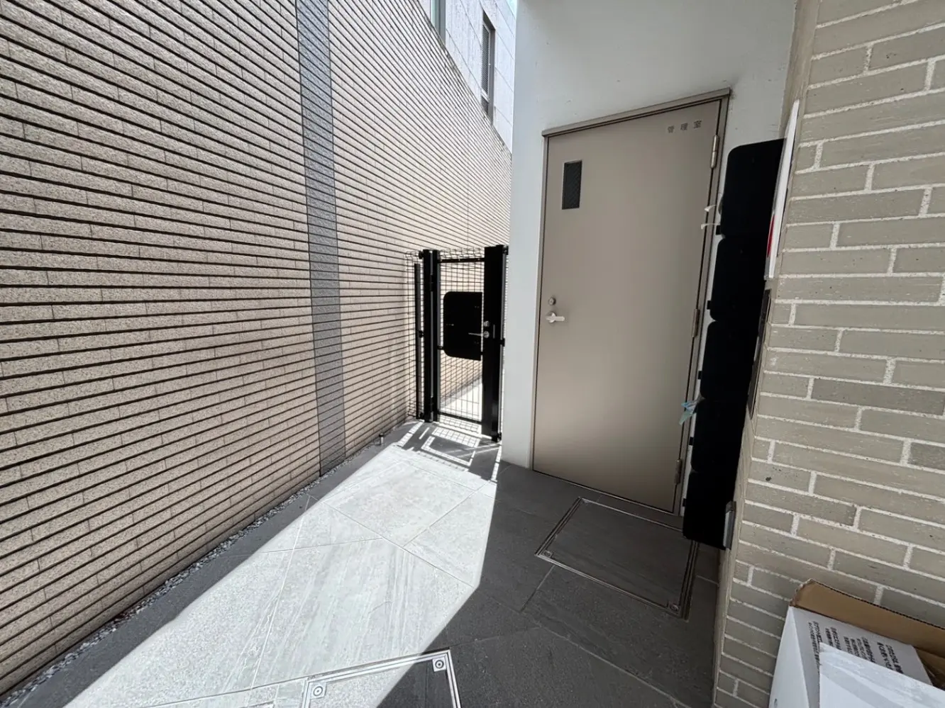 - 壁に囲まれた狭い通路、開いた扉の向こうに郵便受けと施錠可能なゲートがある入口の風景
