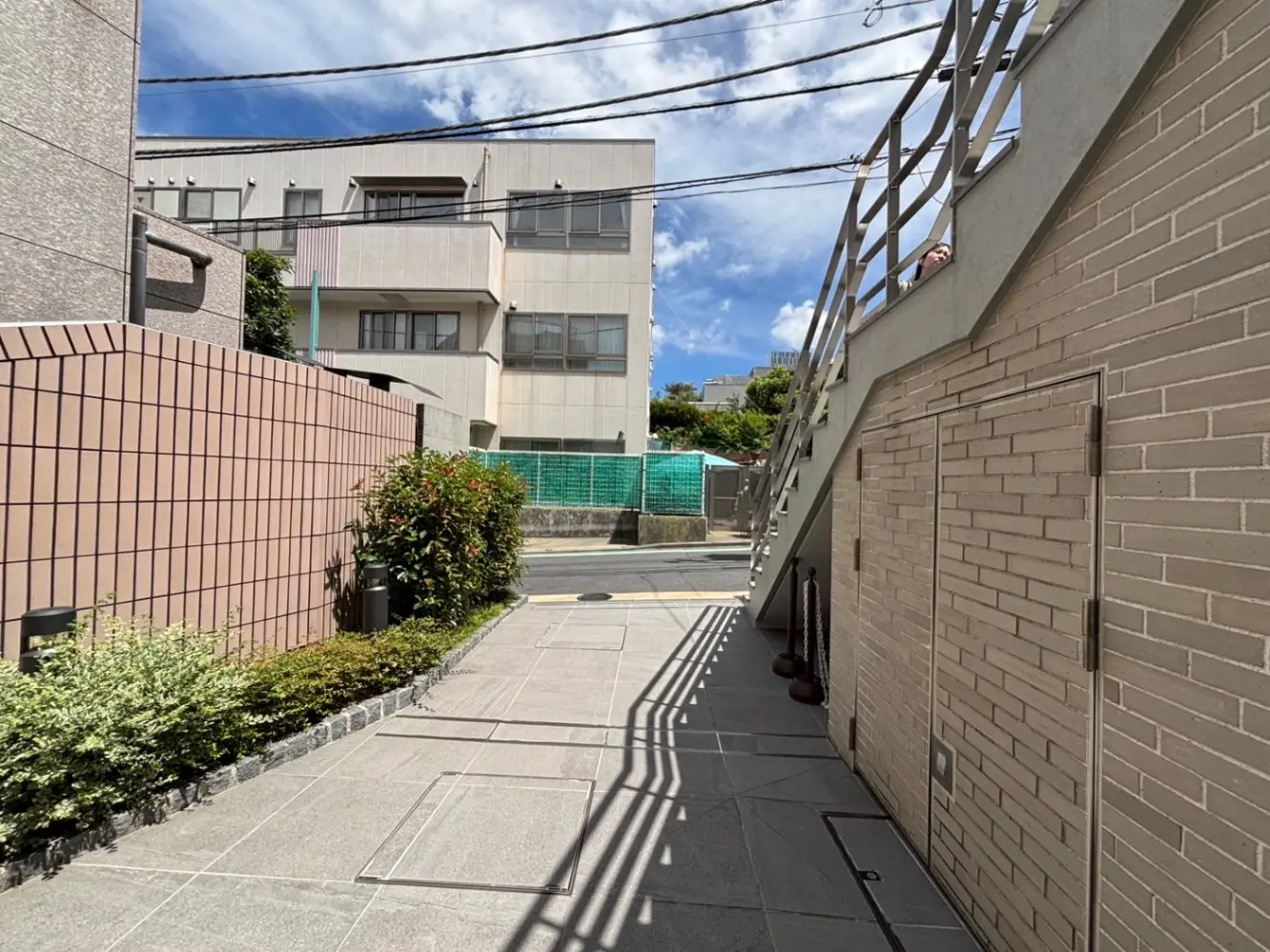 - 高陽光が差し込むアプローチ、近代的な壁面と庭木、階段が見える外観の住宅シーン